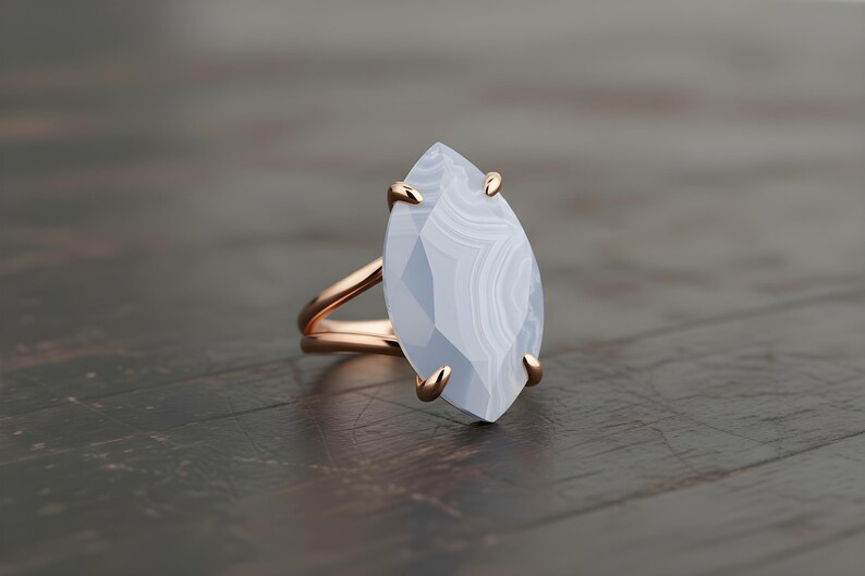 May include: A rose gold ring featuring a large, light blue gemstone with a unique, elongated shape. The gemstone has a marbled appearance. The ring is set on a dark wooden surface.