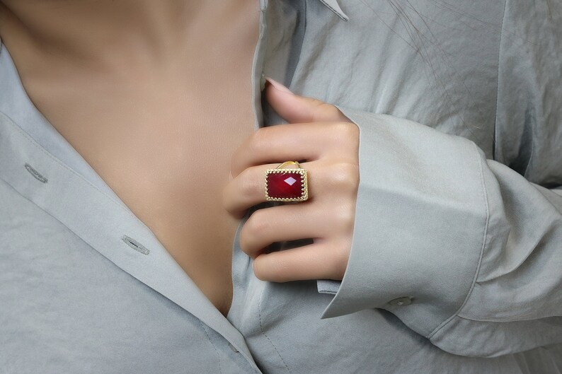May include: A gold-toned ring with a large, rectangular, faceted red gemstone. The ring has a detailed, textured border. The image shows the ring on a person's finger, with a gray shirt in the background.