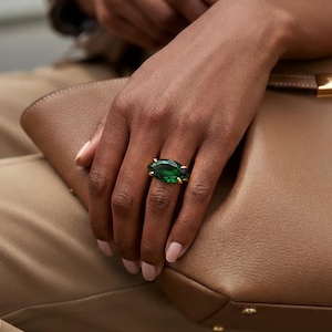 Puede incluir: Un anillo de oro con una gran gema verde esmeralda de forma ovalada. El anillo está en un dedo, junto a un bolso de mano de cuero marrón claro. El fondo incluye ropa de color beige.