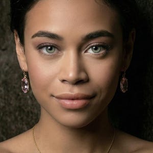 May include: A close-up portrait of a person wearing pink gemstone earrings. The earrings are oval-shaped and dangle from gold-colored hooks. The person has light skin, green eyes, and a subtle smile. A gold necklace is visible.