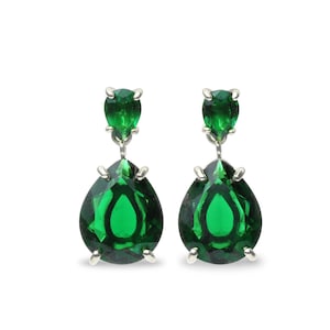 Op de afbeelding: Een paar oorhangers met heldergroene, peervormige edelstenen. Elke oorbel heeft een kleinere, ronde groene steen boven de druppel. De oorbellen zijn gezet in een zilverkleurig metaal, tegen een effen witte achtergrond.