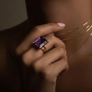 May include: A gold ring featuring a large, rectangular, purple gemstone. The ring is on a hand, with several delicate gold necklaces visible. The image has a warm, soft lighting.