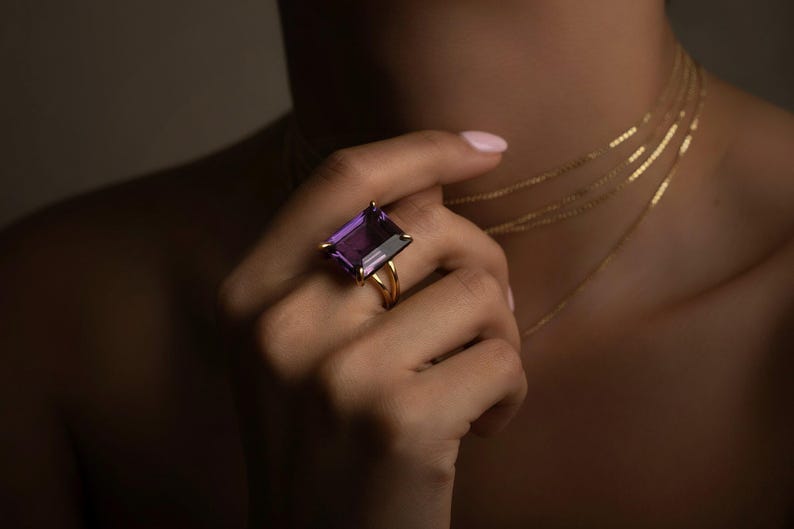 May include: A gold ring featuring a large, rectangular, purple gemstone. The ring is on a hand, with several delicate gold necklaces visible. The image has a warm, soft lighting.