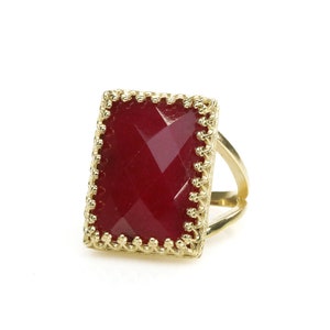 May include: A gold-colored ring with a rectangular, faceted, deep red gemstone. The gemstone is set within a decorative, scalloped gold frame. The ring's band has a split design.
