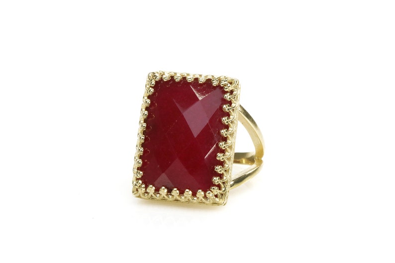May include: A gold-colored ring with a rectangular, faceted, deep red gemstone. The gemstone is set within a decorative, scalloped gold frame. The ring's band has a split design.