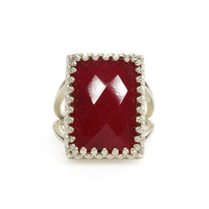 May include: A silver ring with a large, rectangular, faceted, deep red gemstone. The gemstone is set in a decorative, scalloped bezel. The ring has a split shank design, adding visual interest to the piece. The ring is set against a white background.