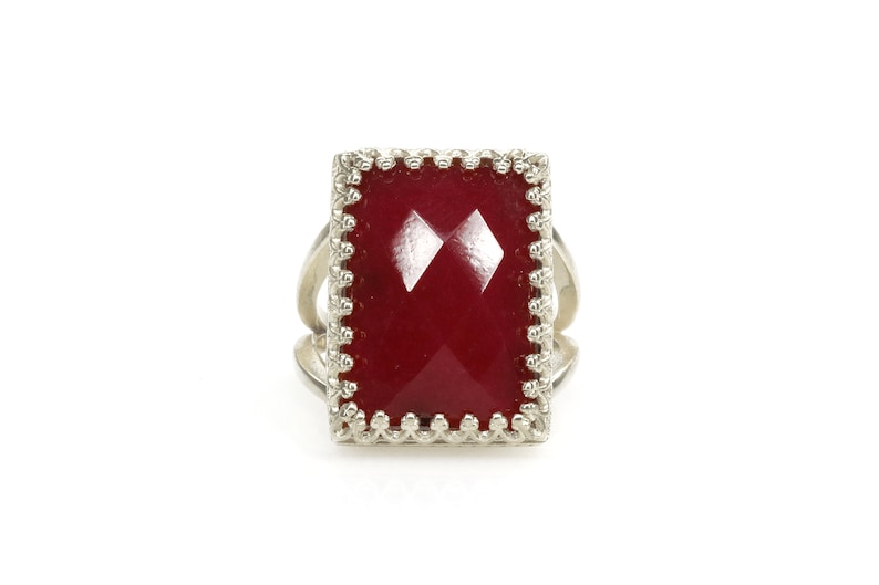 May include: A silver ring with a large, rectangular, faceted, deep red gemstone. The gemstone is set in a decorative, scalloped bezel. The ring has a split shank design, adding visual interest to the piece. The ring is set against a white background.
