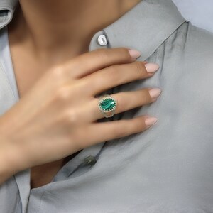 May include: An oval-shaped ring featuring a vibrant green gemstone, set in a silver or white gold band. The gemstone is surrounded by a delicate, textured border, adding to the ring's intricate design. The ring is displayed on a hand.