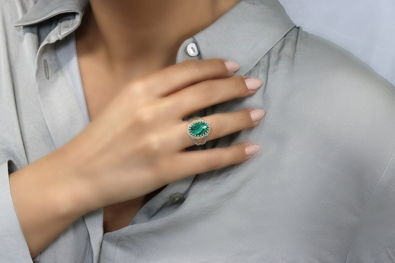 May include: An oval-shaped ring featuring a vibrant green gemstone, set in a silver or white gold band. The gemstone is surrounded by a delicate, textured border, adding to the ring's intricate design. The ring is displayed on a hand.