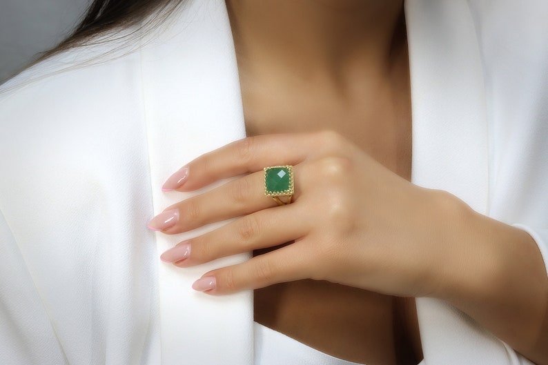 Puede incluir: Un anillo de oro con una gran piedra preciosa verde de corte cuadrado. El anillo tiene una banda de oro texturizada y un borde decorativo. El anillo se muestra en una mano, destacando la joya.