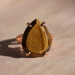 A close-up image of a large, irregularly shaped gemstone, possibly a diamond or a tiger's eye, set in a ring with a metal band.