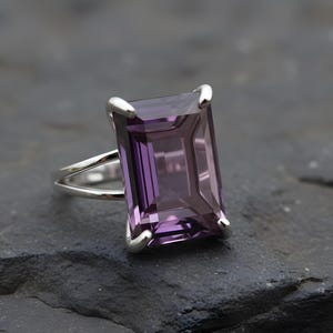 May include: A silver ring featuring a large, rectangular, faceted amethyst gemstone. The amethyst is a deep purple color and is set in a silver band with a split shank design. The ring is displayed on a dark, textured surface.