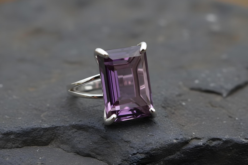 May include: A silver ring featuring a large, rectangular, faceted amethyst gemstone. The amethyst is a deep purple color and is set in a silver band with a split shank design. The ring is displayed on a dark, textured surface.
