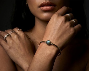 May include: A gold bangle bracelet featuring a single round, iridescent dark gray gemstone at the center. The wrist is adorned with this minimalist piece, while the fingers display multiple thin gold rings with small clear stones.
