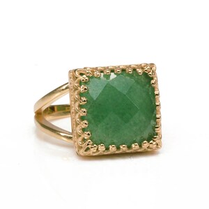 Puede incluir: Un anillo dorado con una piedra preciosa verde cuadrada y facetada. El anillo tiene una banda dividida y un engaste decorativo de estilo filigrana alrededor de la piedra preciosa. El anillo est&aacute; sobre un fondo blanco.