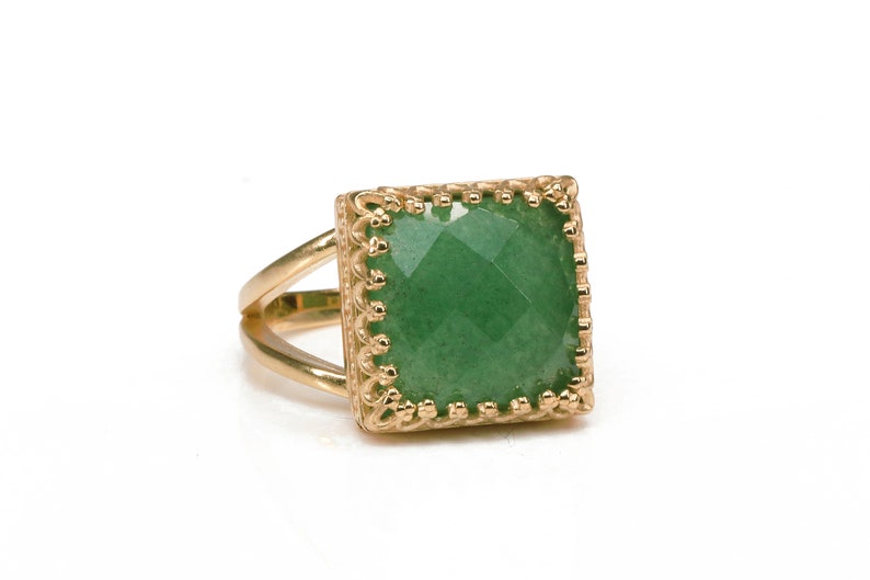 Puede incluir: Un anillo dorado con una piedra preciosa verde cuadrada y facetada. El anillo tiene una banda dividida y un engaste decorativo de estilo filigrana alrededor de la piedra preciosa. El anillo est&aacute; sobre un fondo blanco.