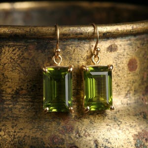 May include: A pair of gold-toned earrings with rectangular, emerald-green gemstones. The earrings are suspended from delicate hooks and set against a textured, antique-looking background.