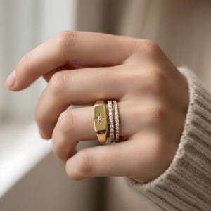 May include: A hand wearing three gold rings. The first is a rectangular signet ring with a starburst design. The second and third rings are thin bands with small, clear stones. The rings are gold in color.