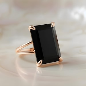 Puede incluir: Un anillo de oro rosa con una gran piedra negra rectangular. El anillo tiene un dise&ntilde;o de ca&ntilde;a dividida y cuatro garras que aseguran la piedra oscura. El anillo est&aacute; sobre un fondo suave y claro.