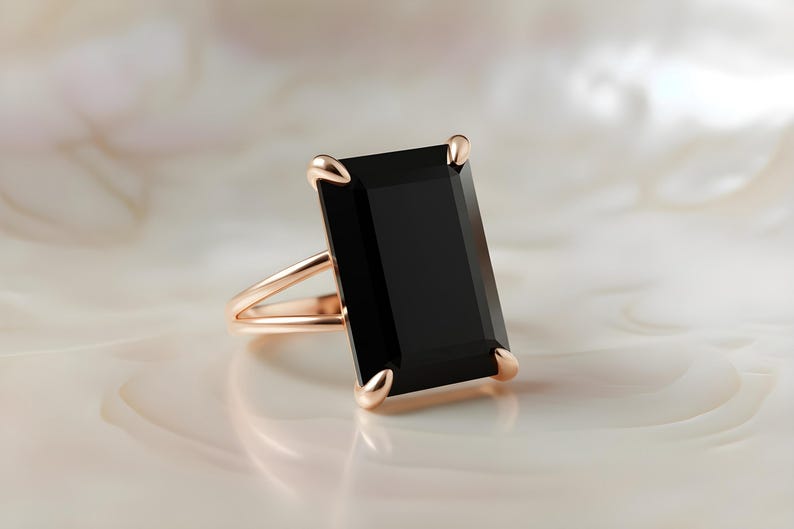 Puede incluir: Un anillo de oro rosa con una gran piedra negra rectangular. El anillo tiene un dise&ntilde;o de ca&ntilde;a dividida y cuatro garras que aseguran la piedra oscura. El anillo est&aacute; sobre un fondo suave y claro.