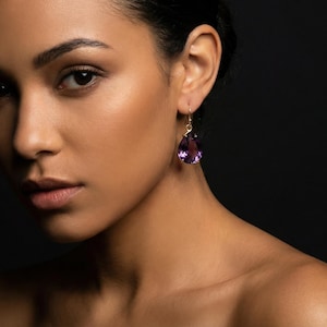 May include: A close-up shot of a person wearing a gold earring with a large, teardrop-shaped purple amethyst. The earring hangs from a gold hook. The background is a dark, solid color.