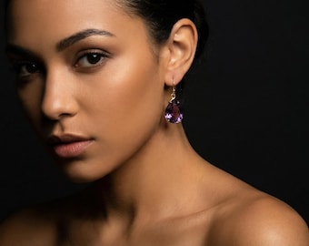 May include: A close-up shot of a person wearing a gold earring with a large, teardrop-shaped purple amethyst. The earring hangs from a gold hook. The background is a dark, solid color.