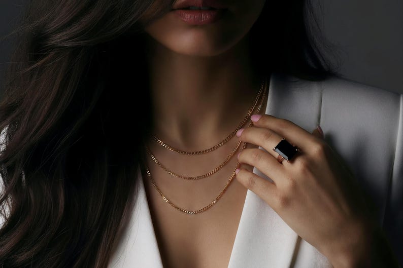 Puede incluir: Un collar de cadena dorada con tres hilos se lleva sobre una blazer blanca. Un gran anillo negro rectangular est&aacute; en la mano. La imagen es un primer plano del cuello y la mano.
