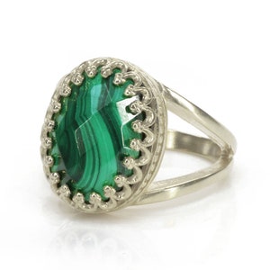 May include: Silver ring featuring an oval malachite stone with a vibrant green pattern. The stone is set in a decorative, scalloped bezel. The ring has a split shank design, adding a modern touch to the jewelry.