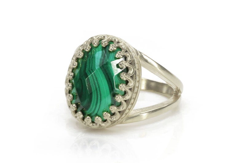 May include: Silver ring featuring an oval malachite stone with a vibrant green pattern. The stone is set in a decorative, scalloped bezel. The ring has a split shank design, adding a modern touch to the jewelry.