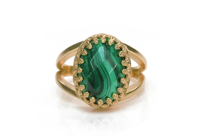 May include: Gold-toned ring featuring an oval malachite stone with a decorative gold border. The ring has a split shank design. The malachite stone displays a vibrant green color with natural veining.