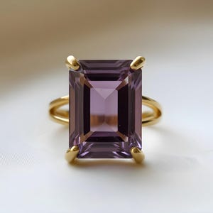 May include: A gold ring featuring a large, rectangular, faceted amethyst gemstone. The amethyst is a deep purple color. The ring has a double band and four gold prongs securing the gemstone.