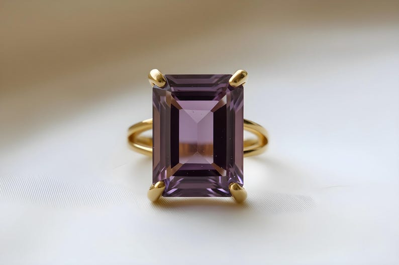 May include: A gold ring featuring a large, rectangular, faceted amethyst gemstone. The amethyst is a deep purple color. The ring has a double band and four gold prongs securing the gemstone.