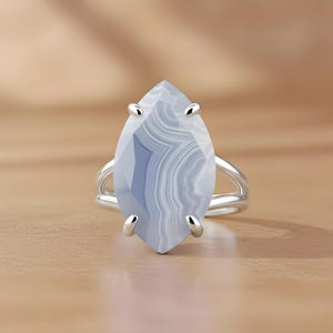 May include: A silver ring featuring a large, marquise-cut gemstone with a blue and white banded pattern. The ring has a split shank design and is set against a light brown surface.