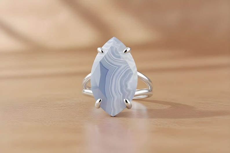 May include: A silver ring featuring a large, marquise-cut gemstone with a blue and white banded pattern. The ring has a split shank design and is set against a light brown surface.