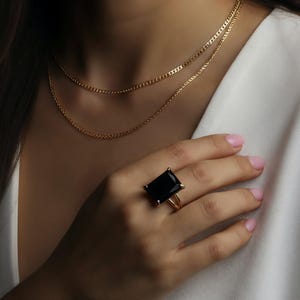Puede incluir: Un collar de oro con dos hilos y un anillo de oro con una piedra negra rectangular grande. El anillo est&aacute; en el dedo de una persona, que lleva una blazer blanca.