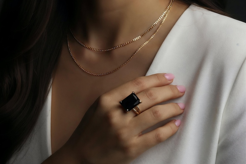 Puede incluir: Un collar de oro con dos hilos y un anillo de oro con una piedra negra rectangular grande. El anillo est&aacute; en el dedo de una persona, que lleva una blazer blanca.