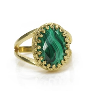 May include: A gold-colored ring with a split shank design. The ring features a large, oval-shaped malachite stone with a vibrant green color and unique patterns. The stone is set in a decorative, scalloped bezel.