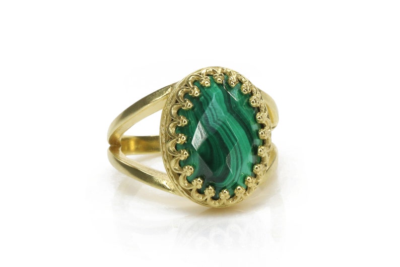 May include: A gold-colored ring with a split shank design. The ring features a large, oval-shaped malachite stone with a vibrant green color and unique patterns. The stone is set in a decorative, scalloped bezel.