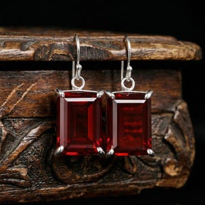 May include: A pair of rectangular, deep red gemstone earrings with silver-colored hooks. The earrings are set in a silver-colored metal and are displayed against a dark, carved wooden surface.