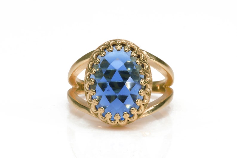 K&ouml;nnte beinhalten: Ein goldener Ring mit einem blauen, ovalen Edelstein, der in einem dekorativen Goldrahmen gefasst ist.