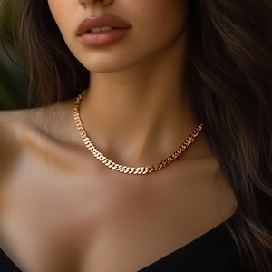 May include: A gold-colored curb chain necklace. The necklace is made of interlocking links and sits close to the neck. The background is blurred, focusing attention on the jewelry.