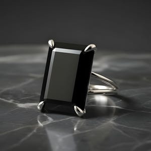 Puede incluir: Un anillo de plata con una gran piedra preciosa negra rectangular. El anillo tiene un dise&ntilde;o de ca&ntilde;a dividida y cuatro garras que aseguran la piedra oscura. El engaste y la banda est&aacute;n pulidos, reflejando la luz sobre un fondo oscuro.