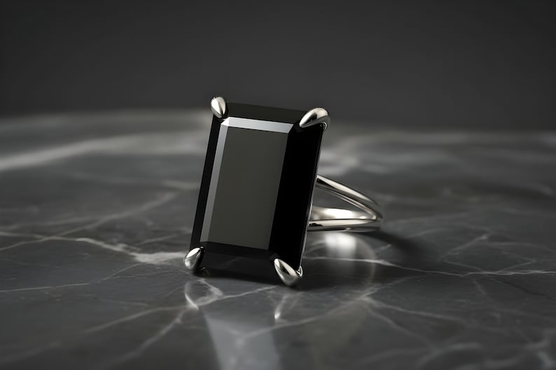 Puede incluir: Un anillo de plata con una gran piedra preciosa negra rectangular. El anillo tiene un dise&ntilde;o de ca&ntilde;a dividida y cuatro garras que aseguran la piedra oscura. El engaste y la banda est&aacute;n pulidos, reflejando la luz sobre un fondo oscuro.