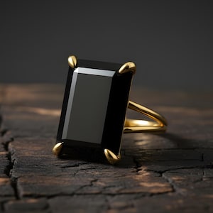 Puede incluir: Un anillo de oro con una gran piedra negra rectangular. El anillo tiene un dise&ntilde;o sencillo con una fina banda de oro y cuatro garras doradas que sujetan la piedra. El anillo est&aacute; sobre una superficie de madera oscura y texturizada.