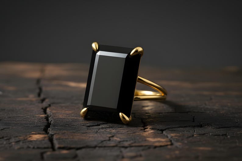 Puede incluir: Un anillo de oro con una gran piedra negra rectangular. El anillo tiene un dise&ntilde;o sencillo con una fina banda de oro y cuatro garras doradas que sujetan la piedra. El anillo est&aacute; sobre una superficie de madera oscura y texturizada.