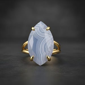 May include: A gold ring with a large, marquise-cut, blue-grey gemstone. The gemstone has a layered, banded pattern. The ring's band is a double-band design, and the gemstone is held in place by four gold prongs.