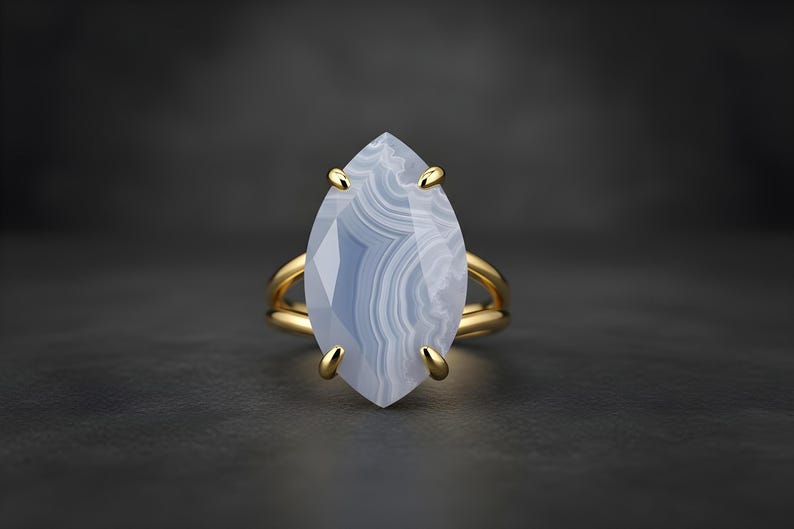 May include: A gold ring with a large, marquise-cut, blue-grey gemstone. The gemstone has a layered, banded pattern. The ring's band is a double-band design, and the gemstone is held in place by four gold prongs.