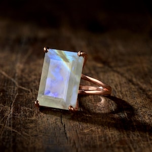 May include: A rose gold ring with a large, rectangular, iridescent gemstone. The gemstone has a pale blue and white color. The ring is set on a dark, textured wooden surface.
