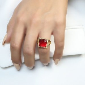 Könnte beinhalten: Ein goldener Ring mit einem quadratischen roten Edelstein, der in einen goldenen Rahmen gefasst ist. Der Ring befindet sich an einem Finger.