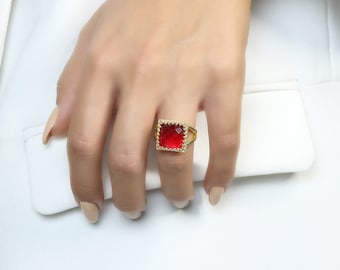 Op de afbeelding: Een gouden ring met een vierkante rode edelsteen gezet in een gouden frame. De ring is op een vinger.
